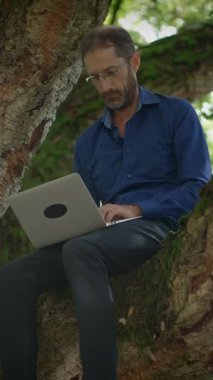 Bir adam rahat bir şekilde dayanıklı bir ağaç dalına oturmuş, özenle laptopuyla dışarıda çalışıyor.