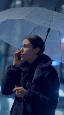 Bir kadın, yağmurlu bir şehir sahnesinde bir şemsiyeyi ustalıkla dengelerken bir telefon görüşmesi yapar.
