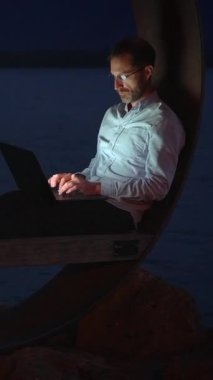 Bir adam geceleri sakin rıhtımda dizüstü bilgisayarıyla çalışıyor. Modern bir uzaktan çalışma tarzı sergiliyor.
