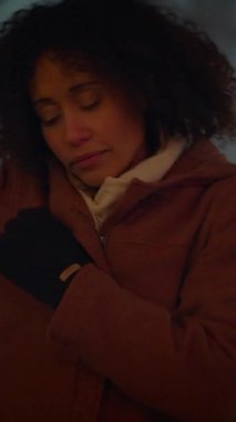 Kıvırcık saçlı bir kadın, rahat bir palto giymiş, şehir hayatını düşünceli bir şekilde yansıtıyor yumuşak, çevresel ışıklar altında.