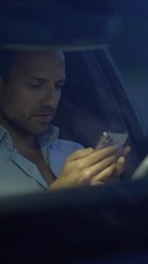 Kendini adamış bir adam, loş bir arabanın içinde rahat bir şekilde otururken akıllı telefonuyla derinden meşguldür.