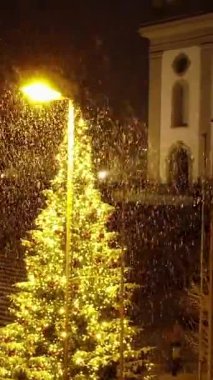 Renkli süslemeleri olan ışıldayan bir Noel ağacı kar tanelerinin altında duruyor ve büyülü bir kış gecesi yaratıyor.