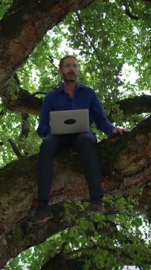 Bir adam rahatça bir ağaca yerleşir, dizüstü bilgisayarıyla çalışır, teknoloji ve doğanın diyarlarını kusursuz bir şekilde harmanlar.