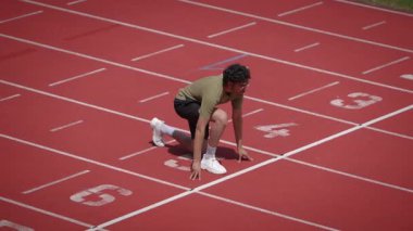 Bir atlet, yoğun bir koşuya tamamen hazır, canlı kırmızı bir pistte başlangıç pozisyonunda duruyor.
