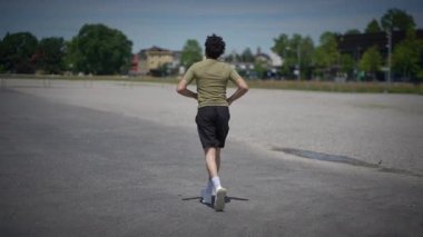 Genç bir insan yemyeşil ve açık gökyüzü ile çevrili boş bir pistte tek başına koşuyor.