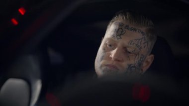 Çarpıcı dövmeli bir adam bir arabanın içinde oturur, benzersiz kişiliğini ve canlı şehir kültürünü yansıtır.