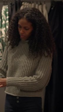 Kıvırcık saçlı genç bir kadın şık, zarif, modern bir ortamda rahat bir süveter giyen eşyaları inceliyor.