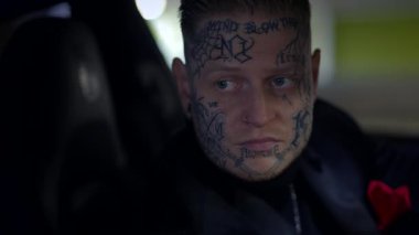 Bir arabada oturan dövmeli bir şahsın görsel olarak çarpıcı portresi, şehir kültürünün özünü mükemmel bir şekilde yakalıyor.