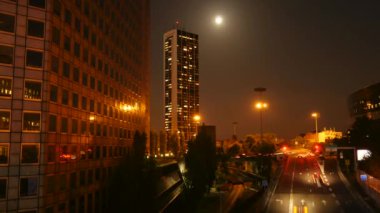 Sokak ışıklarıyla aydınlatılan yüksek binaları ve gökyüzünde parlak ayı gösteren bir gece manzarası..