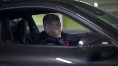 Kamera, bir yeraltı otoparkında park etmiş parlak, koyu renk bir spor arabanın içinde oturan bir sürücüyü yakalıyor.