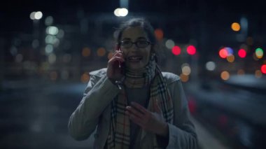Bir kadın, arka plandaki canlı şehir ışıkları tarafından çerçevelenmiş, gece canlı bir telefon görüşmesiyle meşgul oluyor.
