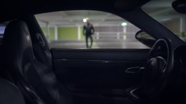 Loş ışıklı bir otoparkta bir araba iç mekan sergileniyor ve uzaklarda tek başına duran bir figür var.
