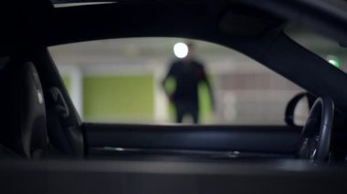 Gölgeli bir figür loş bir otoparkta duruyor, gösterişli bir aracın içinde entrika ve şüphe yaratıyor.
