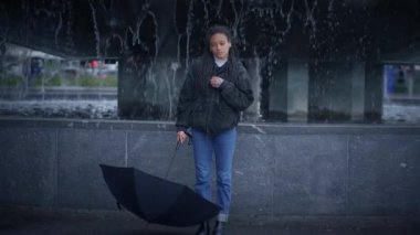 Düşünceli genç bir kadın zarif bir şekilde şemsiyesiyle duruyor, zarif bir şehir çeşmesiyle çerçevelenmiş.