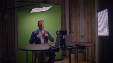 Takım elbiseli bir adam masada oturmuş arkasında yeşil ekran olan önemli bir video kaydına hazırlanıyor.