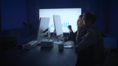 Loş bir ofiste, iki profesyonel geç saatlere kadar çalışır, ekranlardaki verileri analiz eder ve bilgi almak için işbirliği yaparlar.