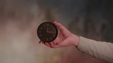 Bir el, renkli bir arkaplan arasında zamanın geçişini sembolize eden antika siyah bir çalar saati tutar..