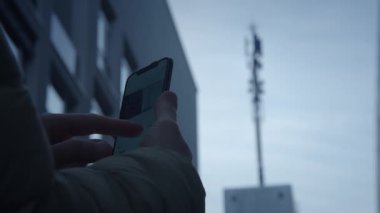 Bir birey, arka planda belirgin bir cep telefonu kulesi olan akıllı bir telefonla meşgul oluyor.