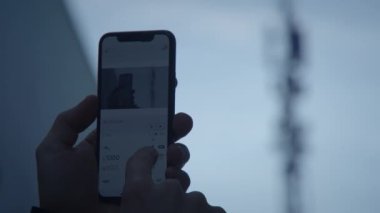 Bir kullanıcı arka planda belirgin bir sinyal kulesi görünürken akıllı telefon aygıtı ile aktif olarak etkileşim içindedir