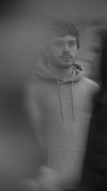 Sıradan bir kapüşonlu giyen genç bir adam dışarıda dikiliyor, düşünceli bir ruh hali içinde ve rahat bir şekilde düşünüyor.