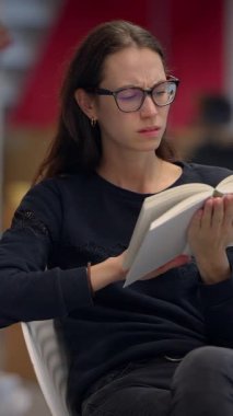 Odaklanmış bir kadın okuyucu, çağdaş bir kütüphane ortamında otururken eğlenceli bir kitapla meşgul oluyor.