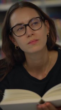 Genç bir kadın, sessiz bir kütüphane ortamında ilginç bir kitap okurken düşüncelerini derinden yansıtıyor.