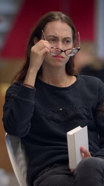 Düşünceli genç bir kadın modern, sanatsal bir ortamda şık bir gözlük ve kitap tutuyor.