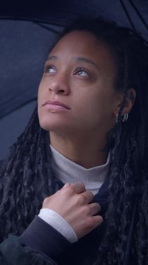 Düşünceli bir kadın, yağmurlu ve huzurlu bir ortamda elinde renkli bir şemsiye tutarken düşünceli bir şekilde yukarı bakar.