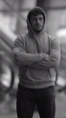 Şık bir kapüşonlu giyen genç bir adam modern bir şehir ortamında kollarını kavuşturarak duruyor.