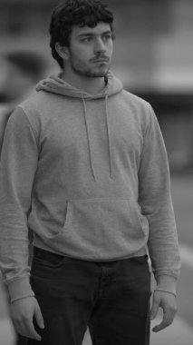 Kapüşonlu genç bir adam düşünceli, canlı, hareketli bir şehir ortamında düşüncelere dalmış duruyor.