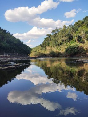 Sıcak ve güneşli bir günde, pek çok ağaç ve kayanın ortasındaki bir nehrin berrak sularına yansıyan bulutlarla dolu mavi gökyüzü.