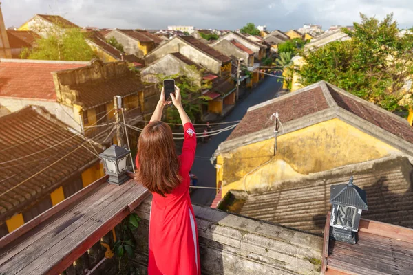 stock image happy woman wearing Ao Dai Vietnamese dress, traveler taking photo by mobile phone on rooftop at Hoi An ancient town in Vietnam. Vietnam and Southeast travel concept