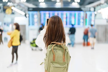 Young woman with backpack looking at the flight time information board  and using smartphone in international airport. Travel, Vacation, trip and Transport concept