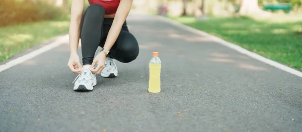 Young athlete woman tying running shoes with Energy Drink water, female runner ready for jogging outside, asian Fitness walking and exercise in the park morning. wellness, wellbeing and sport concepts