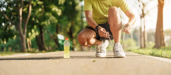 Young athlete man tying running shoes with Energy Drink water, male runner ready for jogging outside, asian Fitness walking and exercise in the park morning. wellness, wellbeing and sport concepts