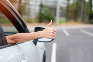 Kadın, yolda araba kullanırken başparmağını kaldırıyor, modern elektrikli otomobilde direksiyonu elle kontrol ediyor. Yolculuk, yolculuk ve güvenlik ulaşım kavramları