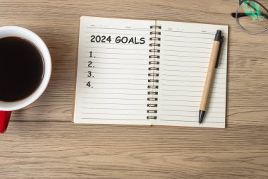 Not defteri, siyah kahve fincanı ve kalemle ahşap masa, üst manzara ve kopyalama alanı olan 2024 GOAL. Mutlu yıllar, karar, yapılacaklar listesi, strateji ve plan konsepti
