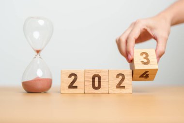 hand flipping block 2023 to 2024 text with hourglass on table. Resolution, time, plan, goal, motivation, reboot, countdown  and New Year holiday concepts clipart