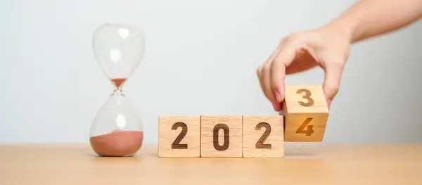 hand flipping block 2023 to 2024 text with hourglass on table. Resolution, time, plan, goal, motivation, reboot, countdown  and New Year holiday concepts