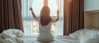 Mutlu kadın uyandıktan sonra yatağa uzanıyor, genç yetişkin kadın kollarını kaldırıyor ve sabahın erken saatlerinde pencereye bakıyor. Rahatlayın ve güzel bir gün geçirin.