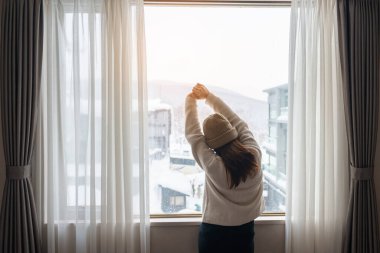 Kazaklı genç bir kadın kış mevsiminde pencereden dışarı bakıyor mutlu kadın kollarını kaldırıyor ve sabah evde ya da dairede uyandıktan sonra esniyor. Yaşam tarzı ve rahatlatıcı kavramlar