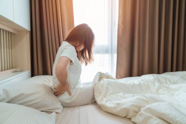 Evde yatakta otururken sırtı ağrıyan bir kadın. Piriforis Sendromu, Düşük Sırt Ağrısı ve Omurga Baskısı nedeniyle uyandıktan sonra kas ağrısı çeken yetişkin bir kadın. Sağlık Medikal Konsepti