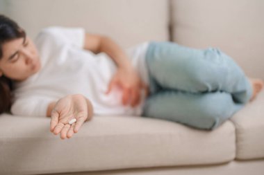 Kadın el ele tutuşup evdeki kanepede ağrı kesici yiyor. Mide ağrısı, ishal, gıda zehirlenmesi, endometriyozis, histerektomi ve regl ağrıları için.