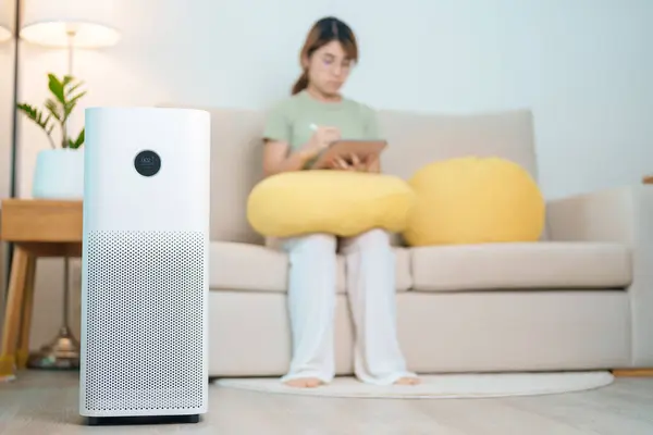 stock image Air Purifier with woman relax and use tablet on sofa. Purification system for filter and cleaning dust PM2.5 HEPA and virus in home. Allergy, Pure air, health, Wellness lifestyle and Air Pollution