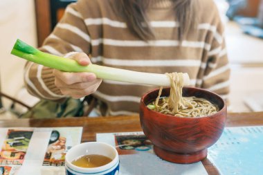 Woman tourist eating Negi Soba or Buckwheat noodles with a raw leek instead of chopsticks, famous soba when visit Ouchi Juku ancient farmer house in Shimogo, Minamiaizu, Fukushima Prefecture, Japan clipart