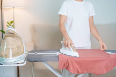 Kadın ütü masasında elektrikli ütü ütülü tişört, akıllı otomatik buhar ütüsü tutuyor. Çamaşır, ev işleri, temizlik, ev temizliği, ev işleri ve günlük rutin konsept.