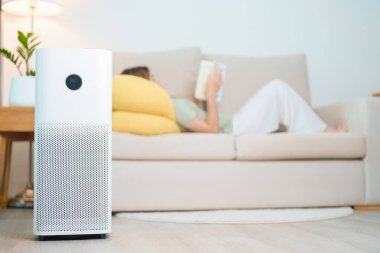 Bir kadınla hava temizleyicisi. Kanepede okuyup rahatlayın. Evde filtre ve temizlik tozu PM2.5 HEPA ve virüs için arıtma sistemi. Alerji, saf hava, sağlık yaşam tarzı, sağlık hayatı ve hava kirliliği.