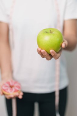 Kadın elini yeşil elma ve çörek ile tutar, kadın fitness meyve ve tatlı sağlıklı ve sağlıksız abur cubur arasında seçim yapar. Diyet kontrolü, kilo kaybı, obezite, yeme tarzı ve beslenme konsepti