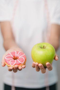 Kadın elini yeşil elma ve çörek ile tutar, kadın fitness meyve ve tatlı sağlıklı ve sağlıksız abur cubur arasında seçim yapar. Diyet kontrolü, kilo kaybı, obezite, yeme tarzı ve beslenme konsepti
