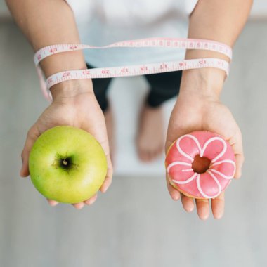 Ağırlık ölçeğinde ve elinde yeşil elma ve çörek olan bir kadın, meyve ve tatlı arasında seçim yapmak sağlıksız ve abur cuburdur. Diyet kontrolü, kilo kaybı, obezite, yeme tarzı ve beslenme